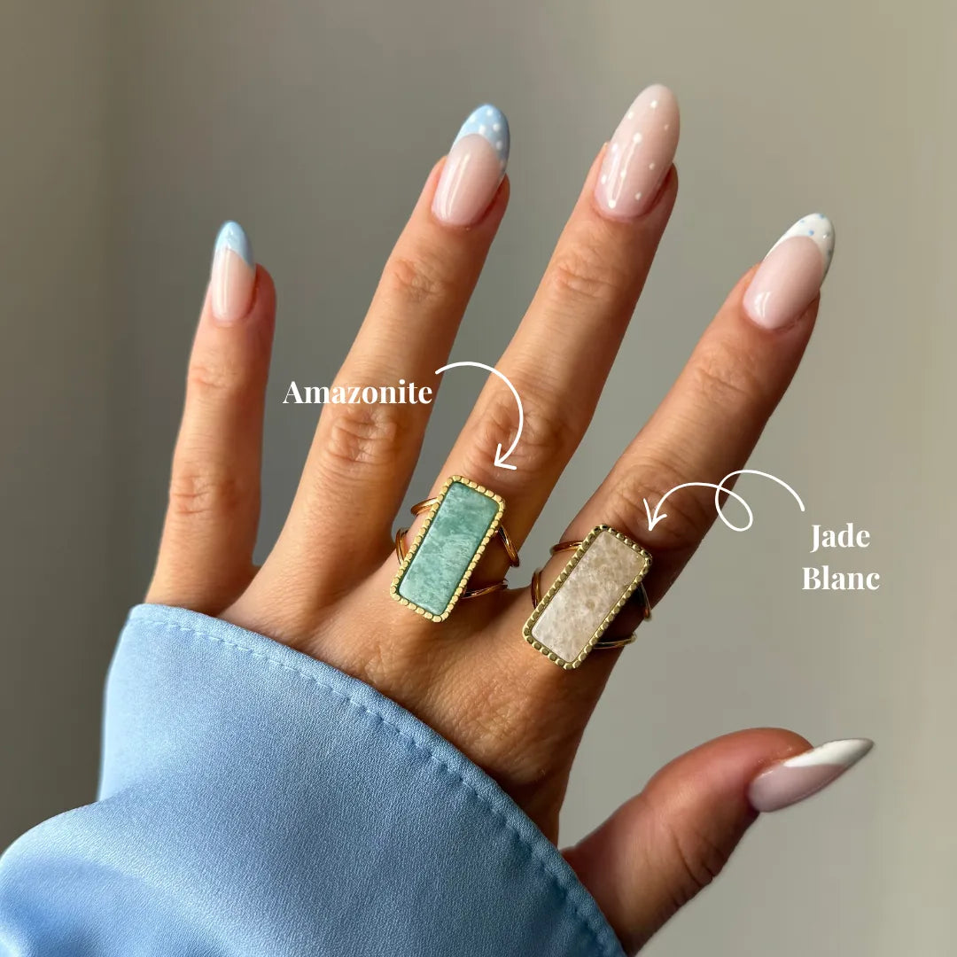 Une main porte deux bagues en or avec pierres rectangulaires : une amazonite verte et un jade blanc crème. Manucure française