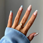 Main sur manche bleue, bague dorée ouverte avec deux pierres ovales opalescentes. Ongles manucure française bleue et à pois.