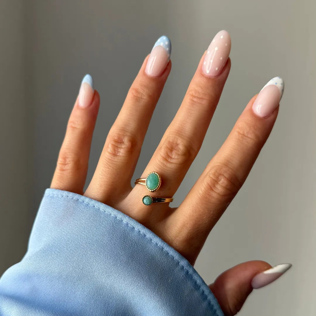 Bague ouverte dorée avec deux pierres vertes, une ovale et une ronde, portée sur le doigt d'une femme aux ongles manucurés.
