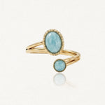Bague dorée ouverte ornée d'une pierre ovale et d'une pierre ronde bleu pâle, avec bordure perlée.