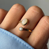 Bague dorée avec deux pierres opalescentes, une ovale et une ronde, portée sur un doigt à côté d'une manche bleue.
