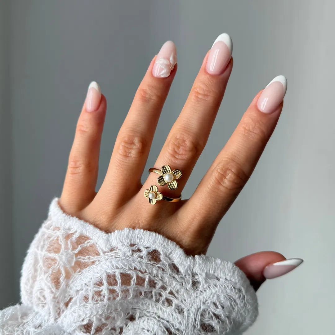 Bague en acier inoxydable doré avec deux motifs fleur et perles blanches, portée sur une main aux ongles manucurés.