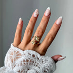 Bague en acier inoxydable doré avec deux motifs fleur et perles blanches, portée sur une main aux ongles manucurés.