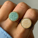 Deux bagues dorées sur une main. Pierres circulaires : l'une bleu-vert veinée, l'autre crème. Anneaux multi-fils et bord