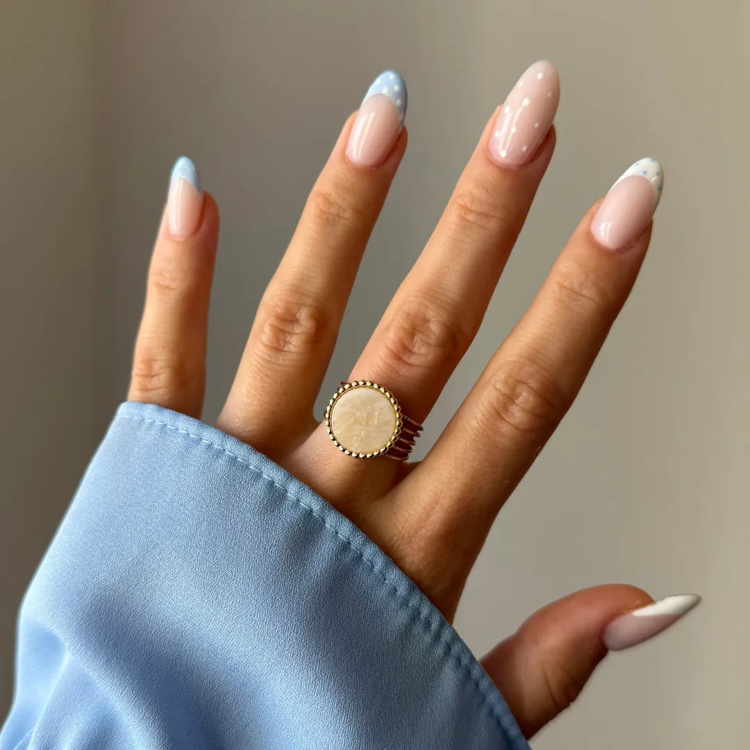 Main montrant une bague dorée avec un plateau rond nacré et un bord perlé. Les ongles sont longs, avec des french manucures