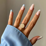 Main montrant une bague dorée avec un plateau rond nacré et un bord perlé. Les ongles sont longs, avec des french manucures