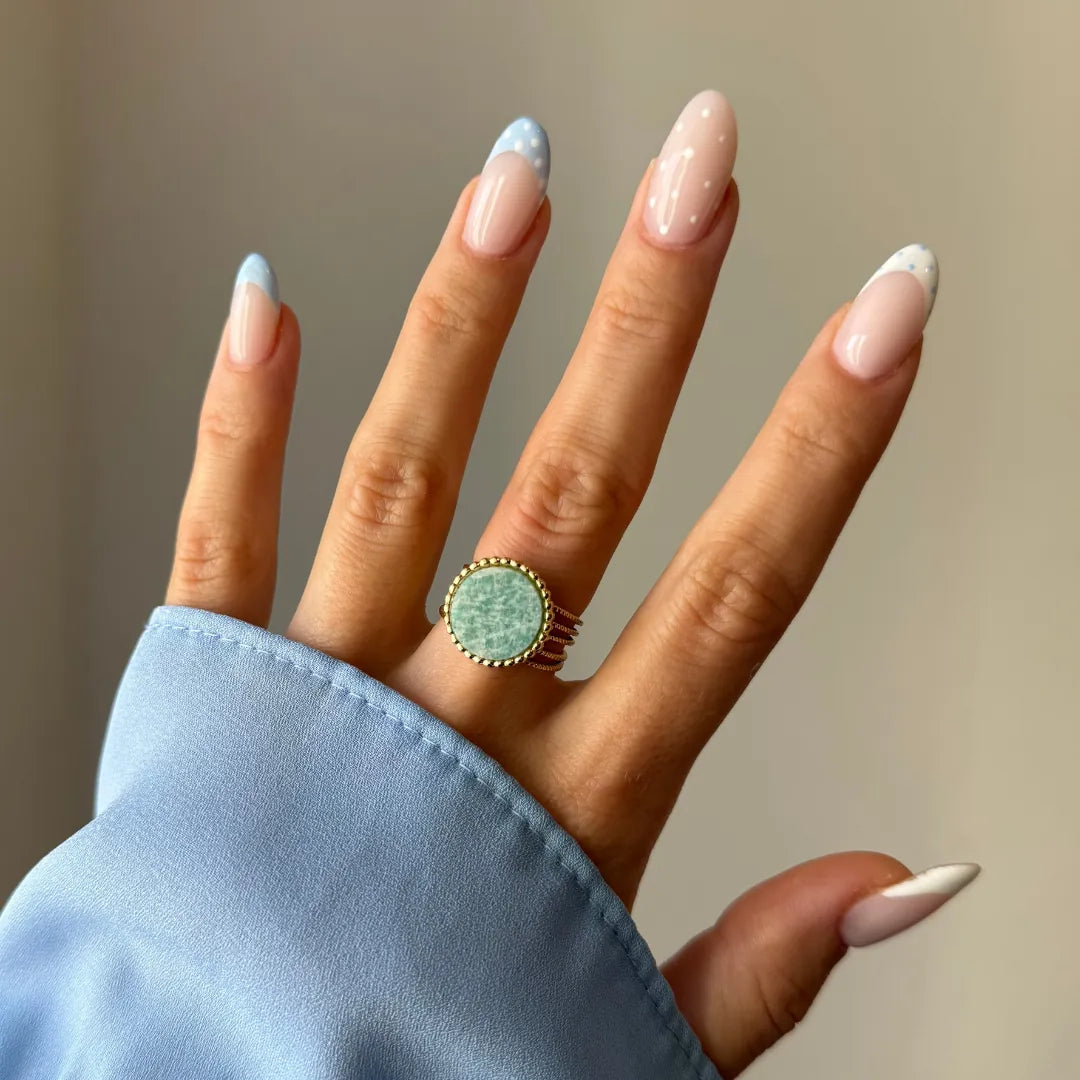 Main avec bague dorée à pierre ronde vert d'eau, bord perlé. Manucure french bleue et points blancs. Manche de chemise bleue.