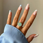 Main avec bague dorée à pierre ronde vert d'eau, bord perlé. Manucure french bleue et points blancs. Manche de chemise bleue.