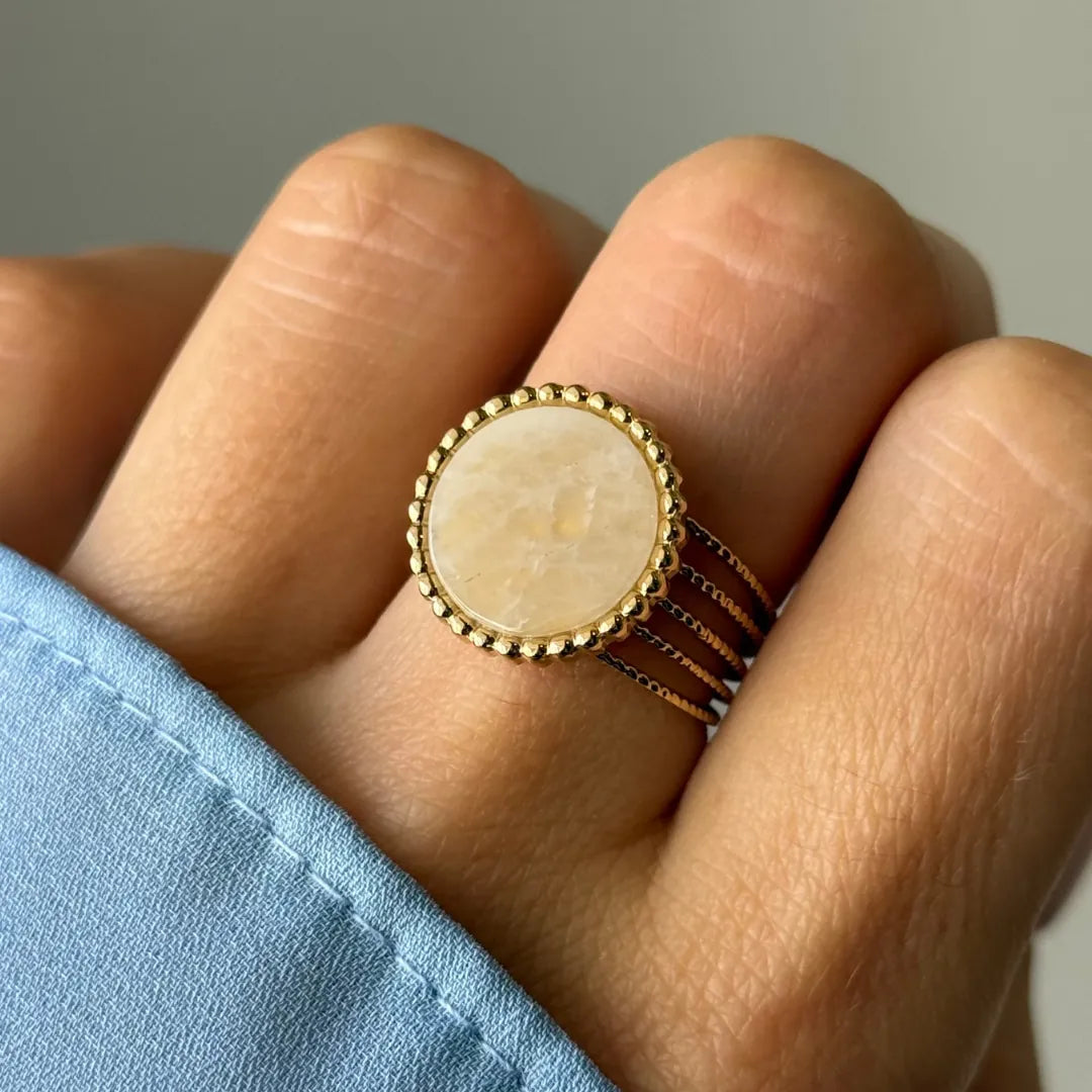 Main portant une bague dorée à plateau rond crème marbré. Le pourtour est perlé et l'anneau est composé de trois fils.