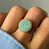 Bague dorée sur un doigt, avec un anneau triple et une pierre ronde vert d'eau texturée, sertie d'un liseré perlé.