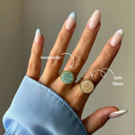 Main montrant deux bagues rondes à bord doré : une Amazonite verte et un Jade Blanc beige. Manucure française bleue.