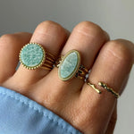 Main portant 3 bagues dorées : une ronde et une ovale avec des pierres vert pâle veinées, et une fine bague ouverte texture