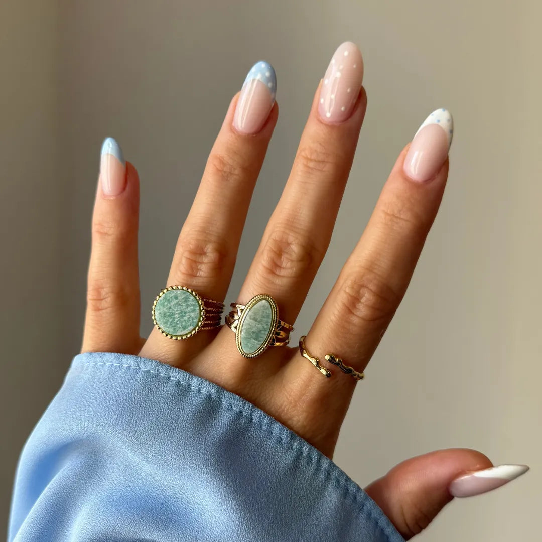 Main de femme portant 3 bagues dorées. Deux ont des pierres rondes/ovales vert pâle, une est ondulée. Manche et ongles bleus.