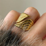 Gros plan sur une bague large dorée aux motifs striés en forme de vagues, portée à l'index.