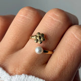 Bague ajustable en acier inoxydable doré sur un doigt, ornée d'une fleur stylisée et d'une perle blanche.
