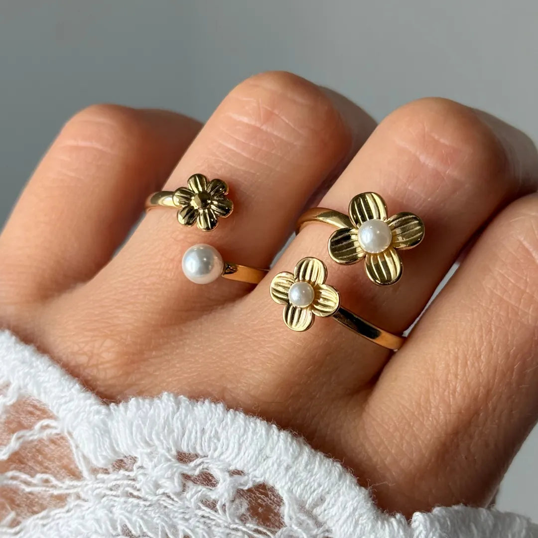 Main portant trois bagues en acier inoxydable doré : une fleur simple, une fleur et perle blanche, deux fleurs et perles