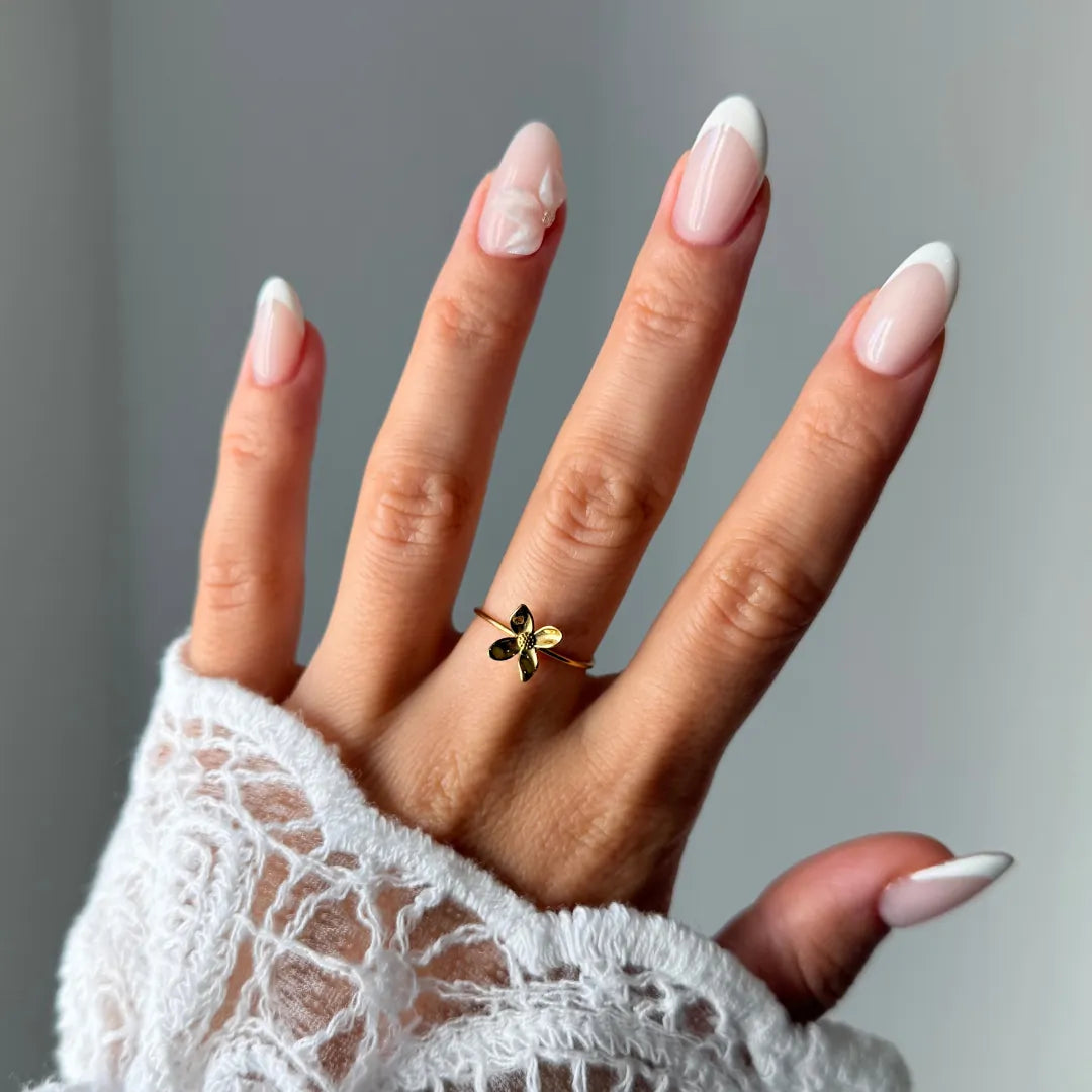 Bague fleur en acier inoxydable doré, portée sur un doigt. La main a des ongles manucurés et une manche en dentelle blanche.