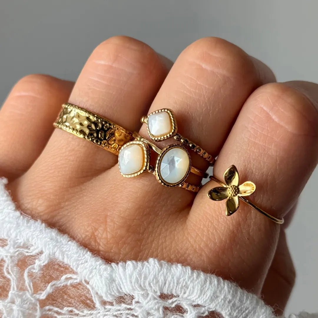 Main ornée d'une bague fleur en acier inoxydable doré et d'autres bagues avec des ornements blancs irisés. Poignet en