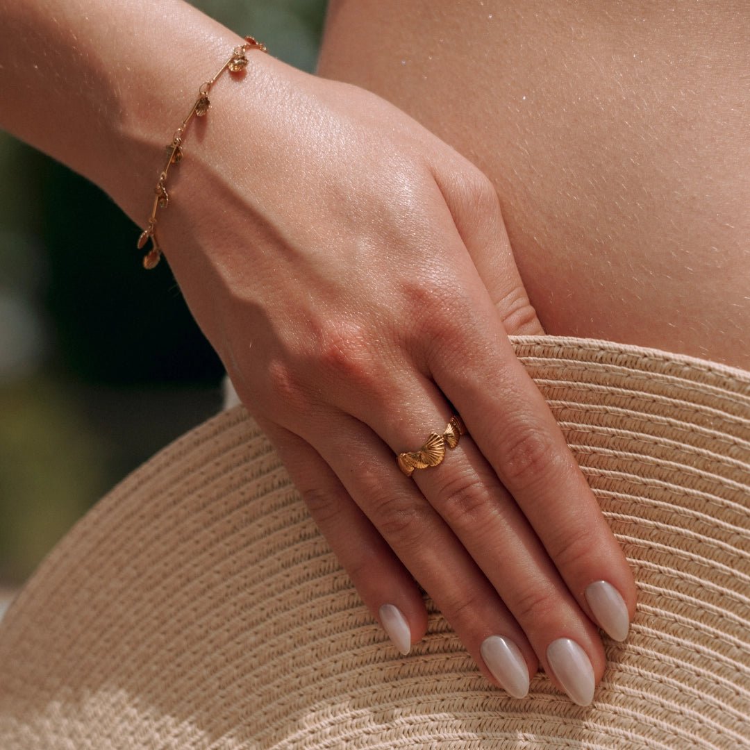 Bague en acier inoxydable doré à motifs coquillages, portée par une main sur un chapeau de paille.