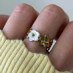 Bague fleurie en acier inoxydable doré avec fleurs en nacre blanche et petites pierres sur doigt, pull jaune.