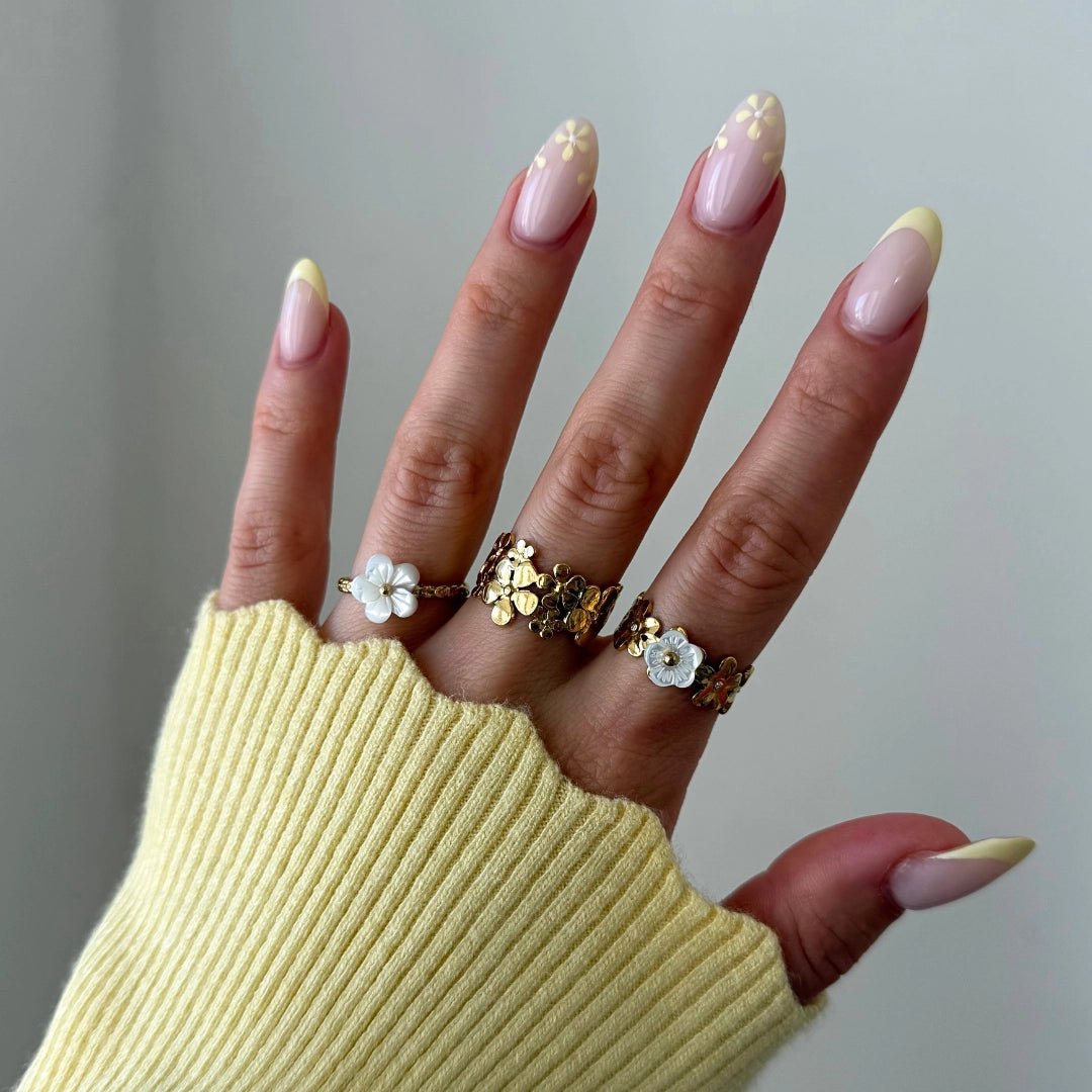 Trois bagues en acier inoxydable doré à motifs floraux (nacre et métal) sur une main en pull jaune.