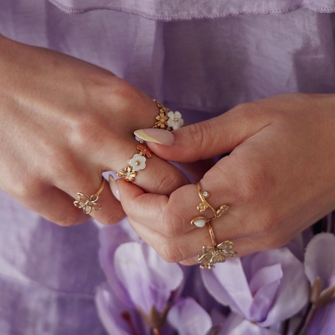 Bagues en acier inoxydable doré, motifs fleurs et perles blanches, portées sur des mains sur fond violet.