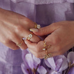 Bagues en acier inoxydable doré, motifs fleurs et perles blanches, portées sur des mains sur fond violet.