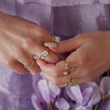 Bagues en acier inoxydable doré, motifs fleurs et perles blanches, portées sur des mains sur fond violet.