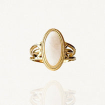 Bague en acier inoxydable doré sertie d'une grande pierre ovale blanche style cabochon sur fond blanc.