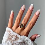 Bague fleur en acier inoxydable doré sur l'annulaire d'une main. La main porte une manucure french, et une manche en dentelle