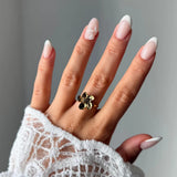 Bague fleur en acier inoxydable doré sur l'annulaire d'une main. La main porte une manucure french, et une manche en dentelle
