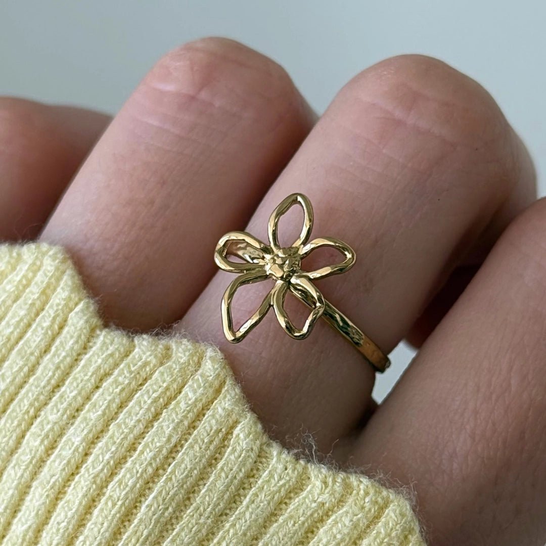 Bague en acier inoxydable dorée, motif fleur ajourée à 5 pétales, portée sur un doigt avec un pull jaune clair.