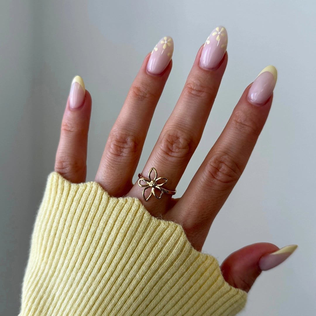 Bague fleur 5 pétales en acier inoxydable doré au doigt, main avec manucure french jaune pastel et pull jaune côtelé.