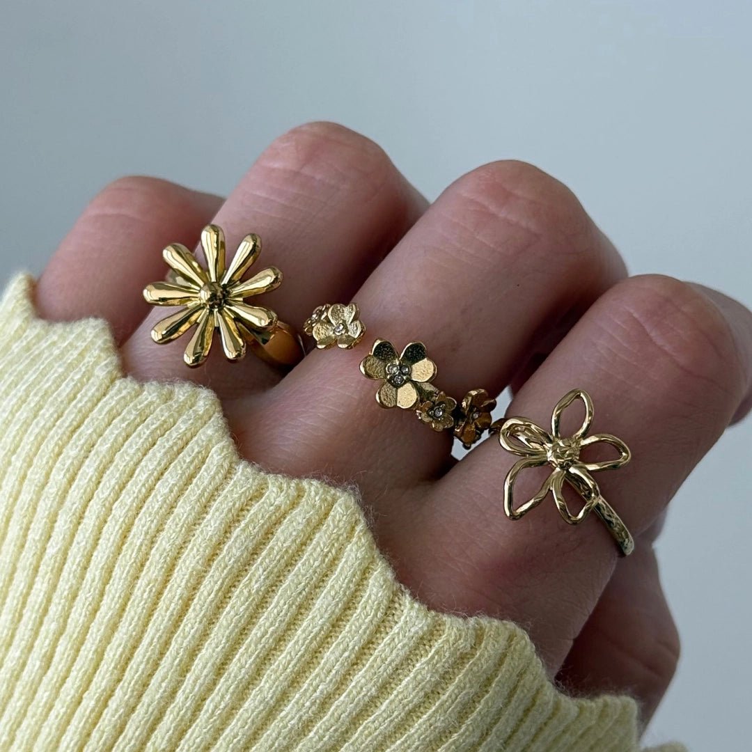 Quatre bagues en acier inoxydable doré à motifs floraux variés sont portées sur une main avec un pull jaune.