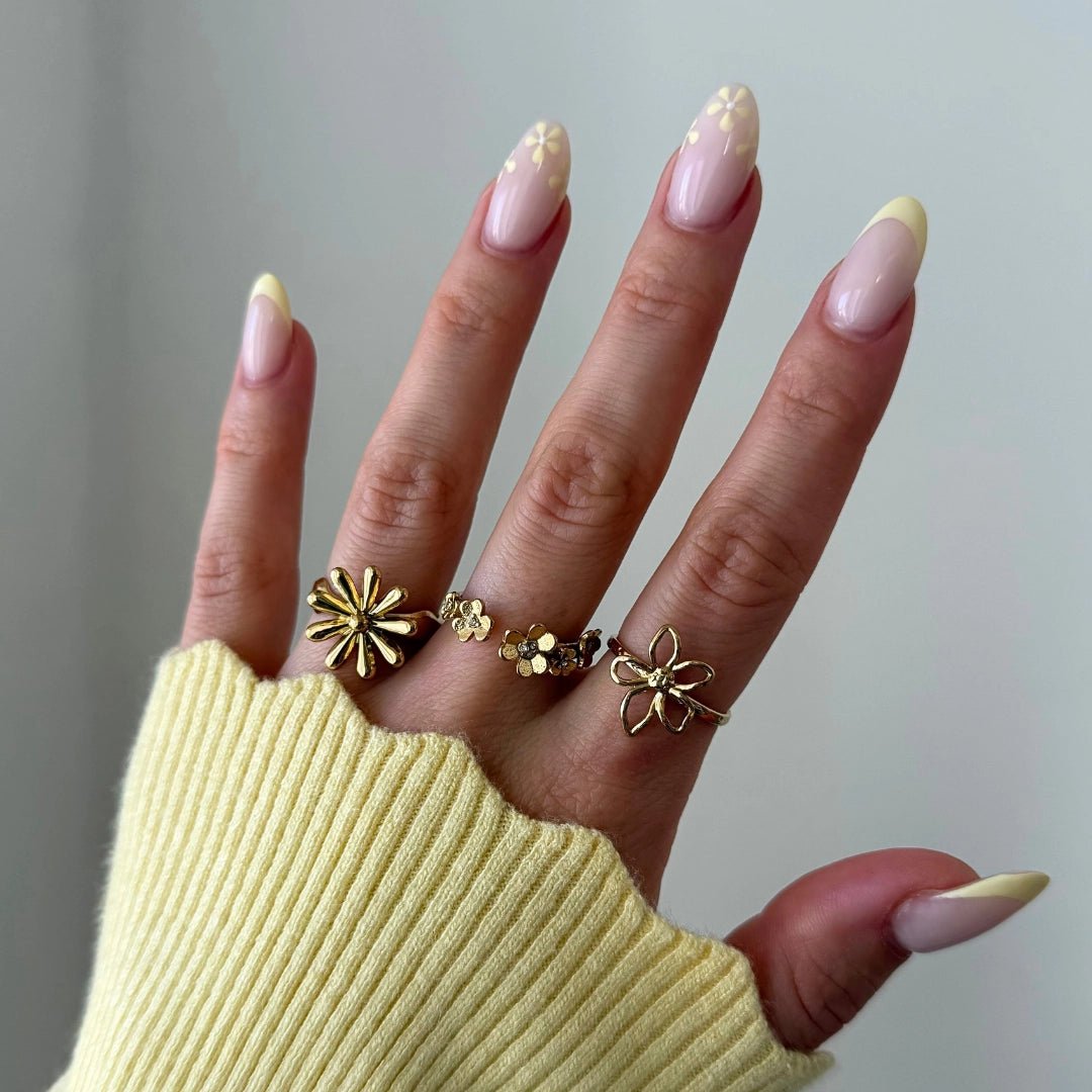 Trois bagues fleur en acier inoxydable doré, portées sur une main féminine avec manucure jaune florale et pull jaune.