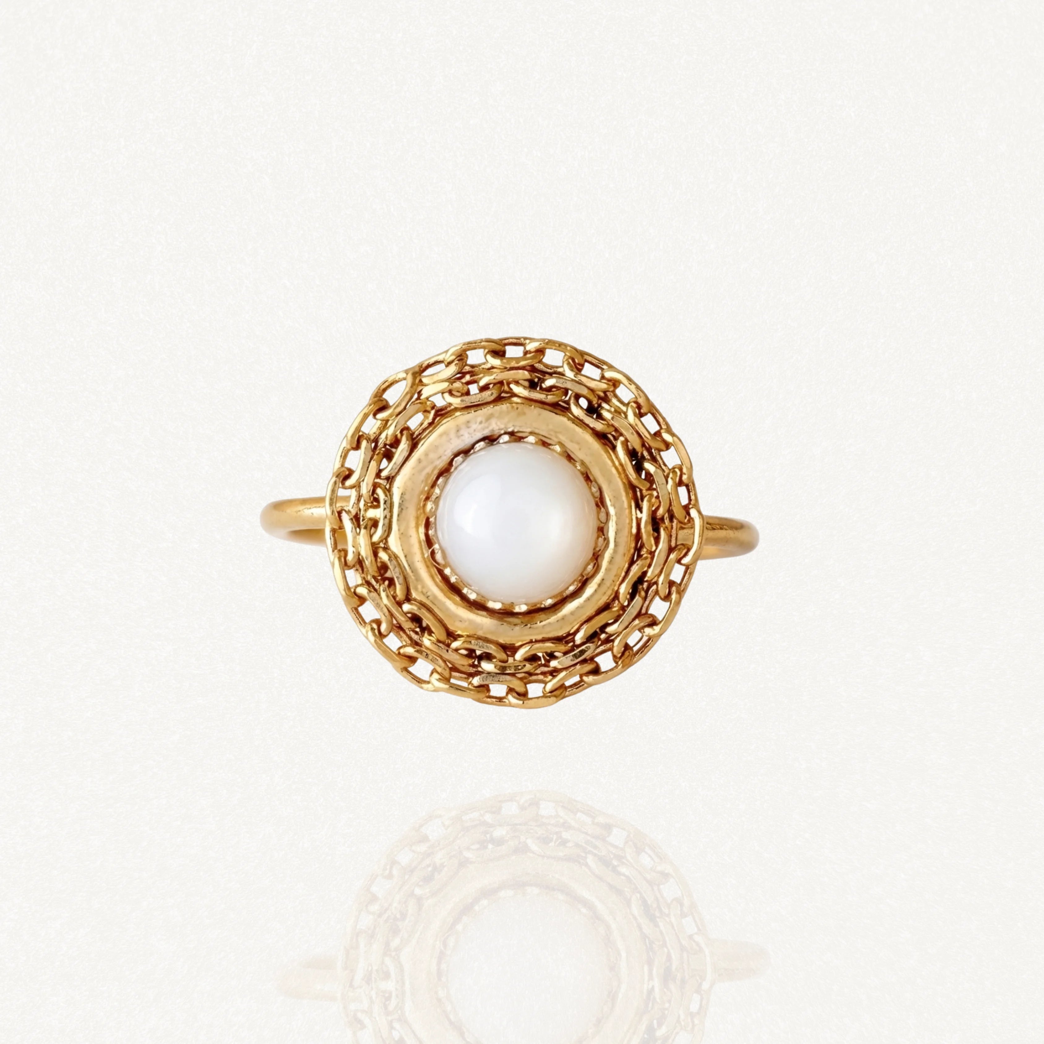 Bague dorée ronde avec une pierre blanche centrale (type nacre) entourée d'un motif maillons de chaîne ajouré sur fond blanc.