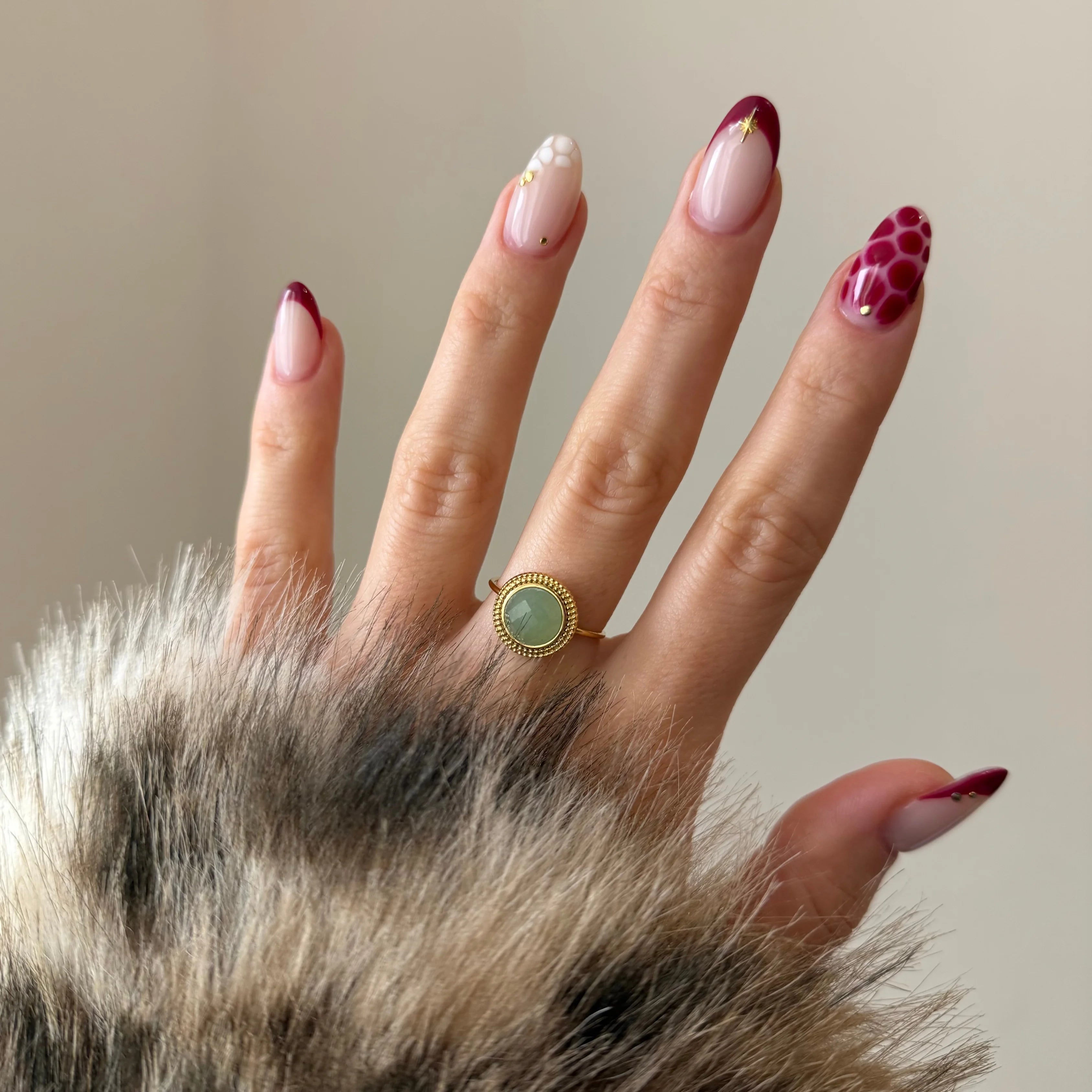 Bague minimaliste dorée avec pierre verte portée à l'annulaire, mise en valeur par un nail art bordeaux.