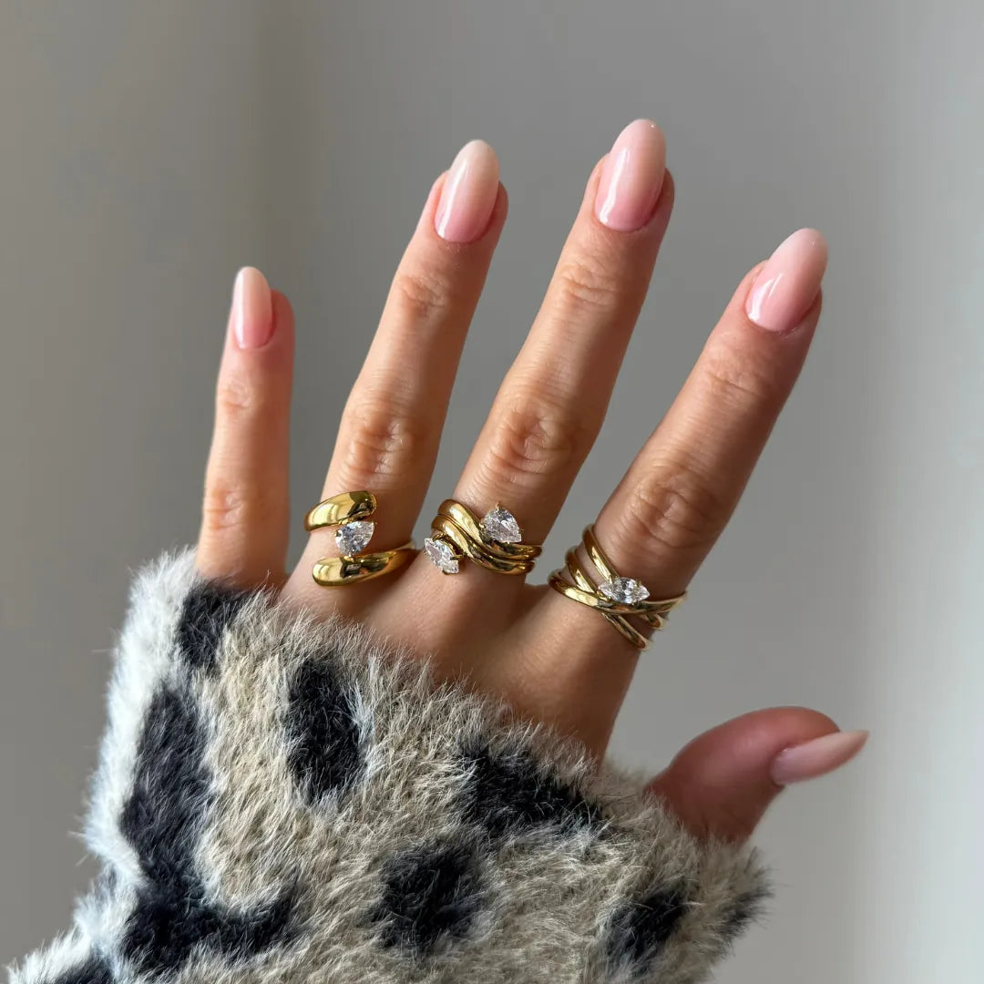 Main aux ongles roses portant une bague multirang entrelacée acier inoxydable doré et zirconium ovale, et deux autres bagues.