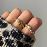 Deux bagues dorées portées: l'une avec perle blanche centrale, l'autre ouverte avec 6 petites perles blanches. Anneaux texturés.