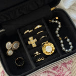 Boîte à bijoux noire ouverte avec divers bijoux dorés: bagues, pendentif croix texturé, trois pièces ovales, et bracelet perles grises.