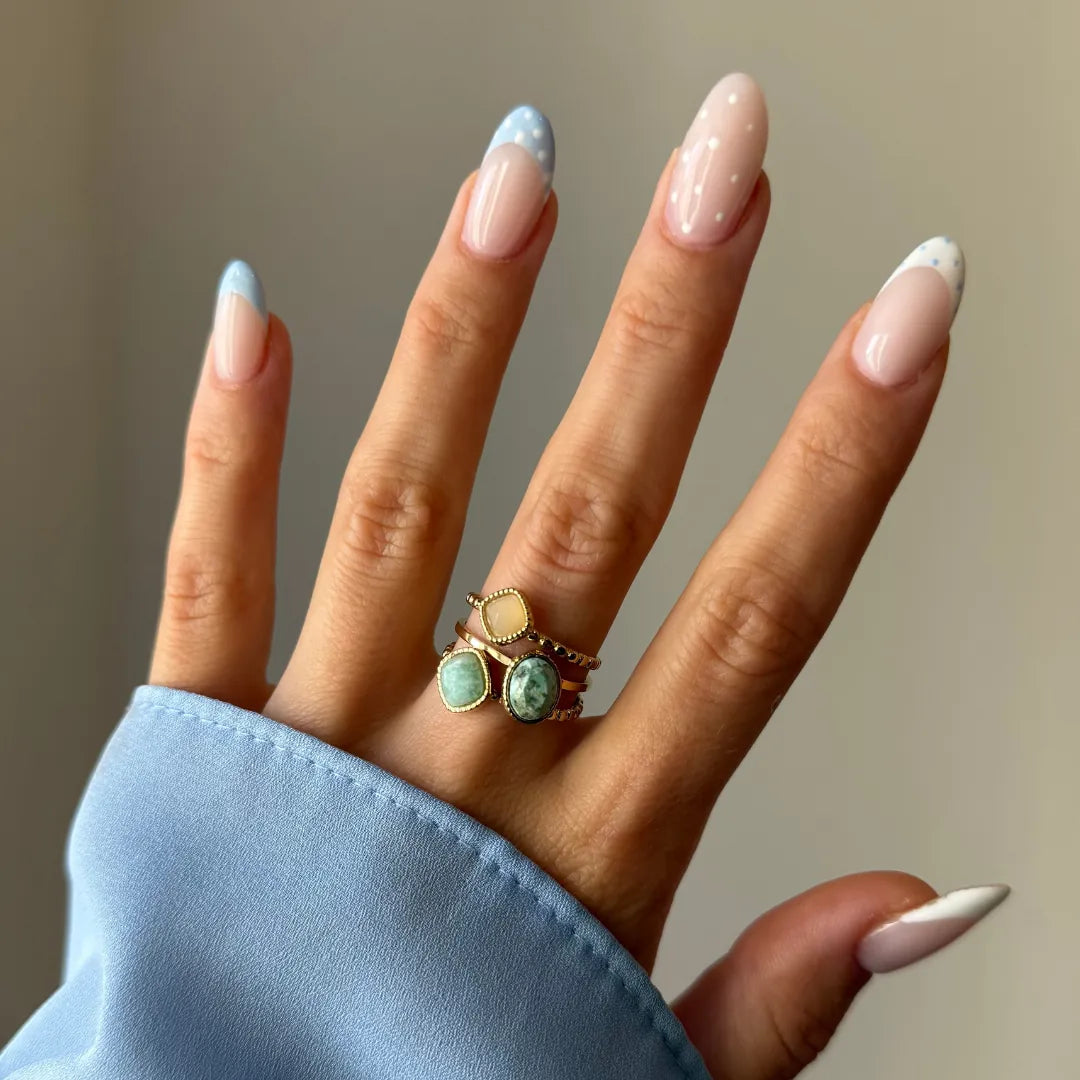 Main manucurée avec des ongles à pois bleus et blancs, portant trois bagues dorées empilées aux pierres variées.