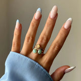 Main manucurée avec des ongles à pois bleus et blancs, portant trois bagues dorées empilées aux pierres variées.