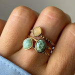 Main avec 4 bagues dorées. Une pierre jaune carrée, une vert clair carrée, une verte marbrée ovale, et une bague perlée.