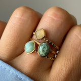 Main avec 4 bagues dorées. Une pierre jaune carrée, une vert clair carrée, une verte marbrée ovale, et une bague perlée.