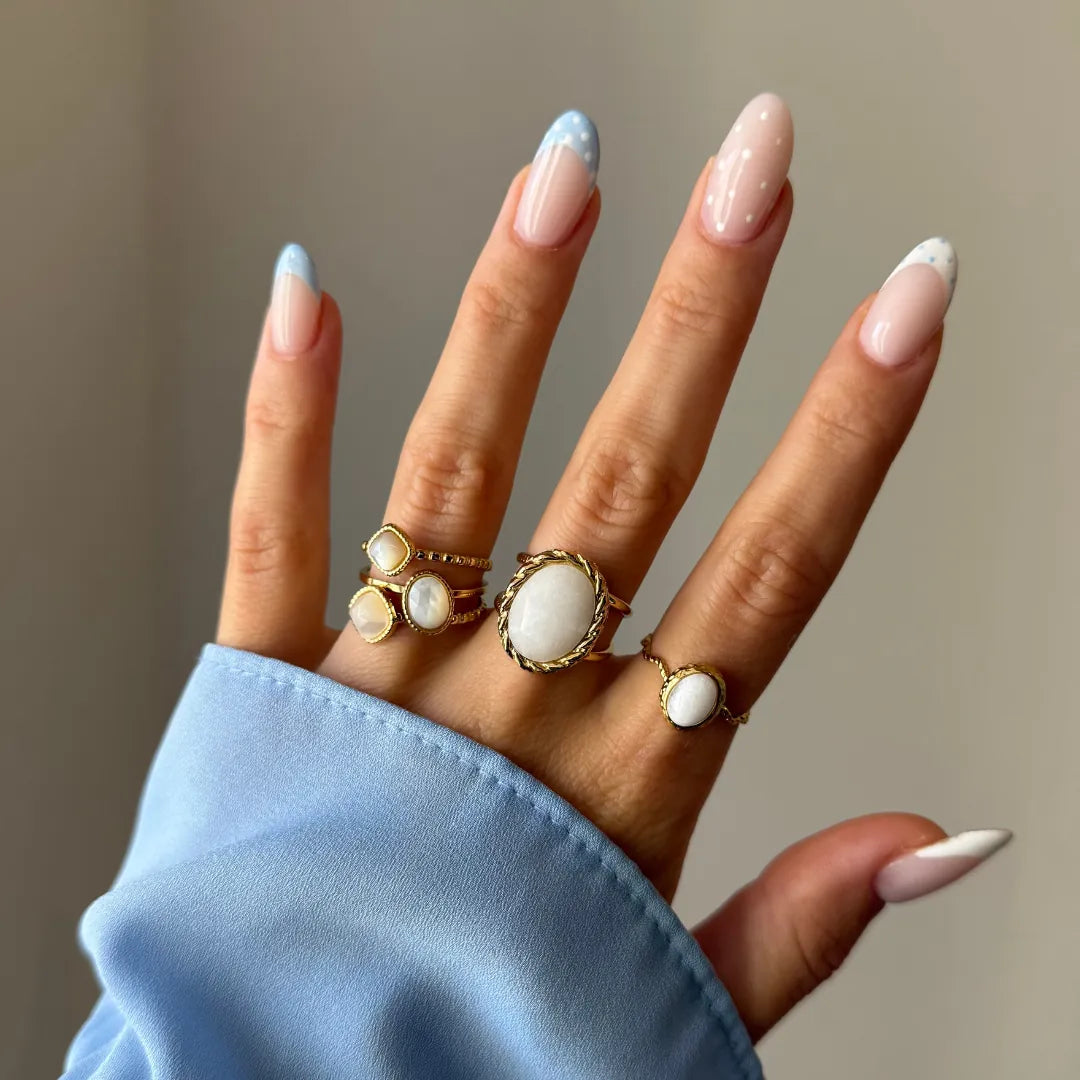 Main portant quatre bagues dorées avec des pierres blanches nacrées, sur des doigts aux ongles bleus et blancs.