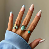5 bagues dorées à pierres (turquoise, verte, crème) sur une main aux ongles manucurés bleu clair à pois.