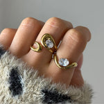 Bague ouverte double goutte en acier inoxydable doré, ornée de deux pierres ovales en zirconium, portée sur un doigt.