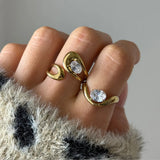 Bague ouverte double goutte en acier inoxydable doré, ornée de deux pierres ovales en zirconium, portée sur un doigt.