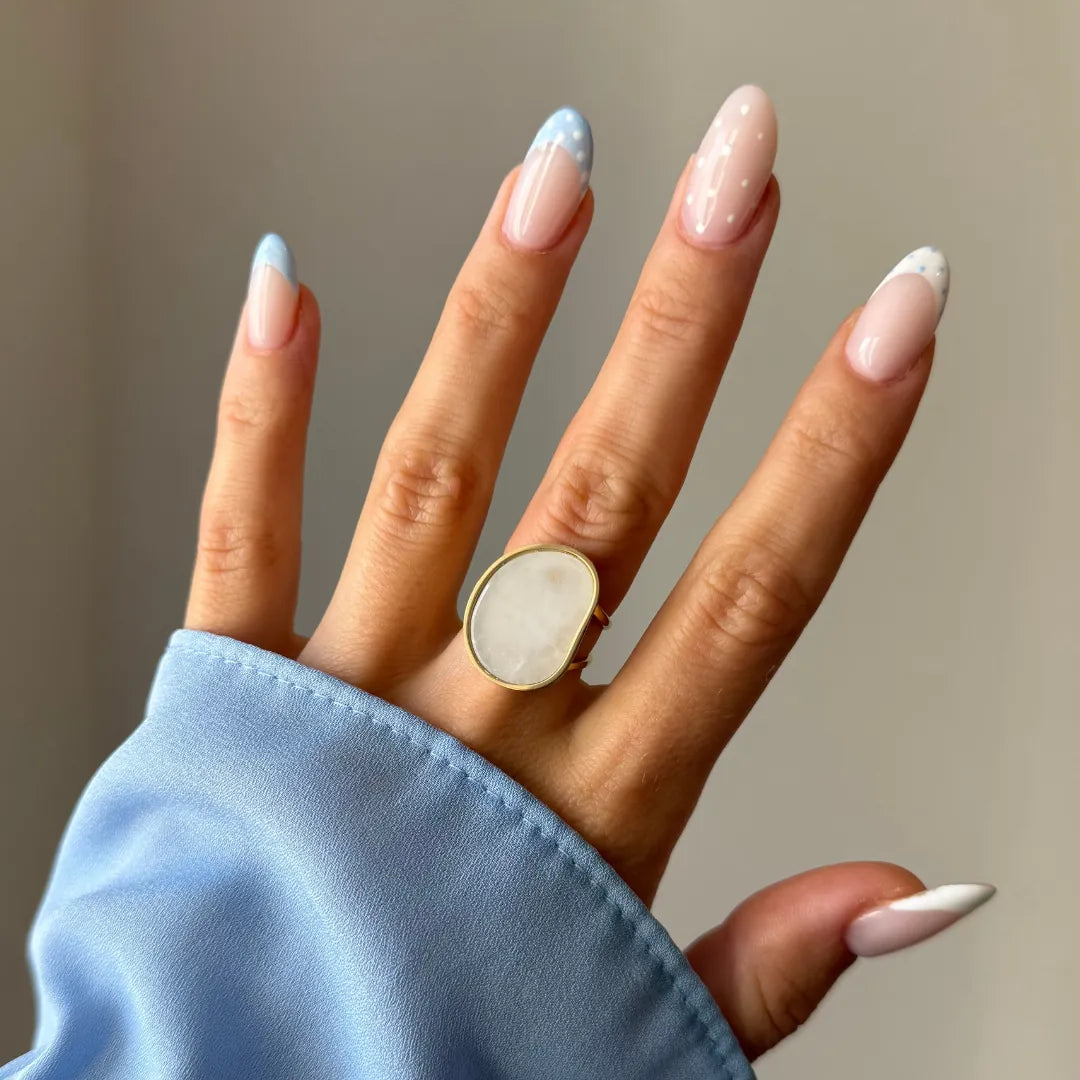 Main avec une bague dorée à pierre laiteuse irrégulière. Ongles longs manucurés avec des pointes bleu ciel à pois et