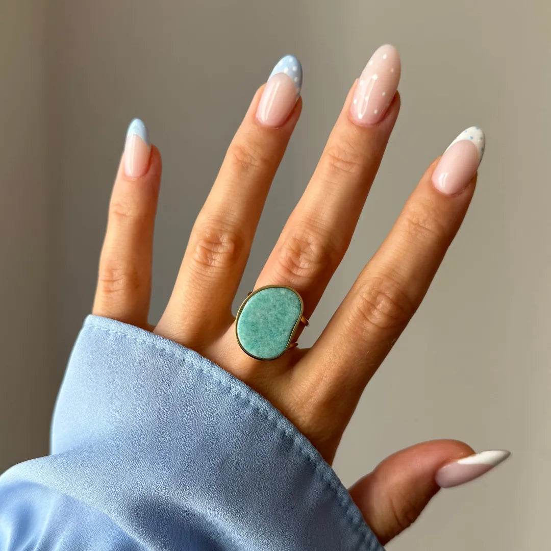 Bague dorée à pierre irrégulière vert d'eau sur le majeur d'une main. Manucure française et manchette de chemise bleue.